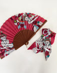 Waaier en bijpassende twilly sjaal van Ottimare met twee Thangka dansers tegen een koraal rode achtergrond met groene accenten. Het houten frame is handgemaakt in Spanje en heeft een goudkleurige ring.