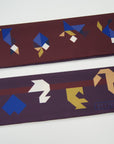 Detail van Ottimare's extra lange Tangram sjaal met twee verschillende kanten. De achtergrond is bordeaux en aan de andere kant Aubergine met een bordeaux streep. De figuren zijn in verschillende kleuren blauwe, camel, goud en ivoor.