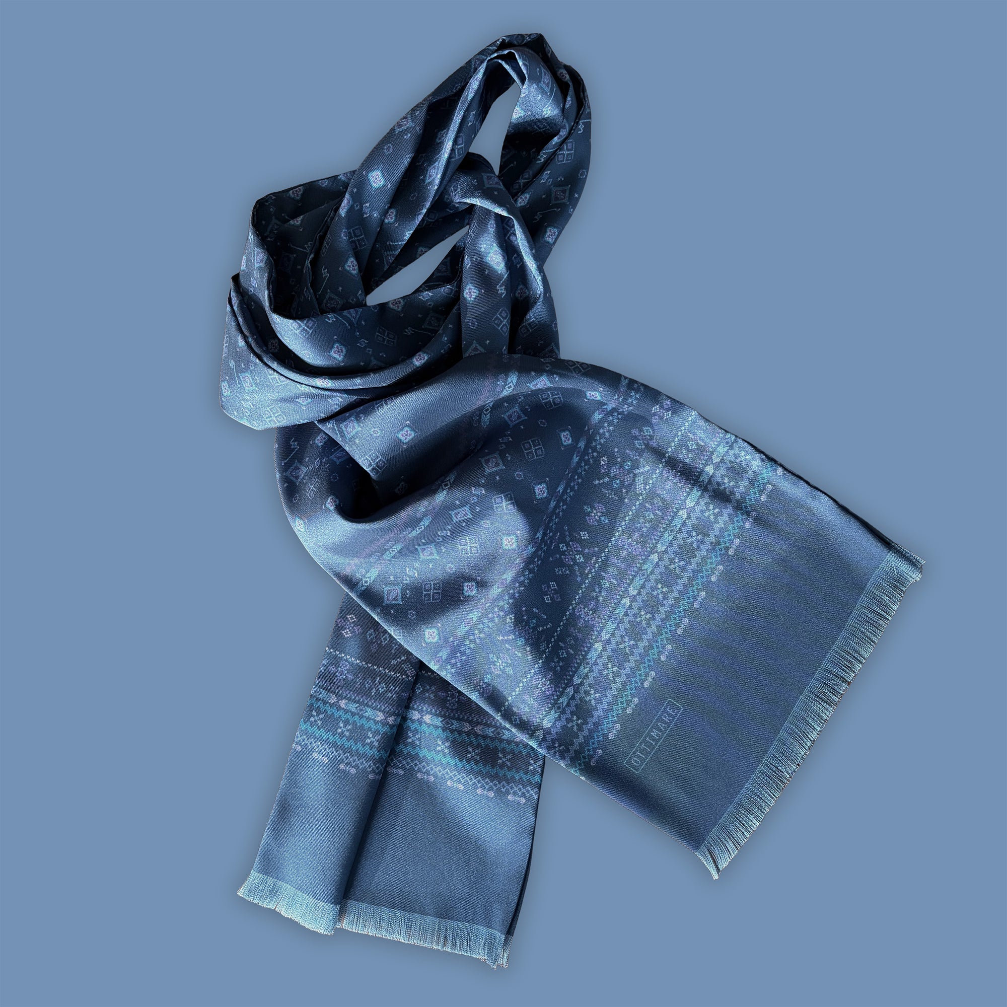 Ottimare's Kahina sjaal geïnspireerd op Berber motieven. Een speciale versie voor Erik, met middelblauw, aqua en paars. De herensjaal met franje die ook door dames gedragen kan worden is handgemaakt in Nederland. De foto heeft een blauwe achtergrond.