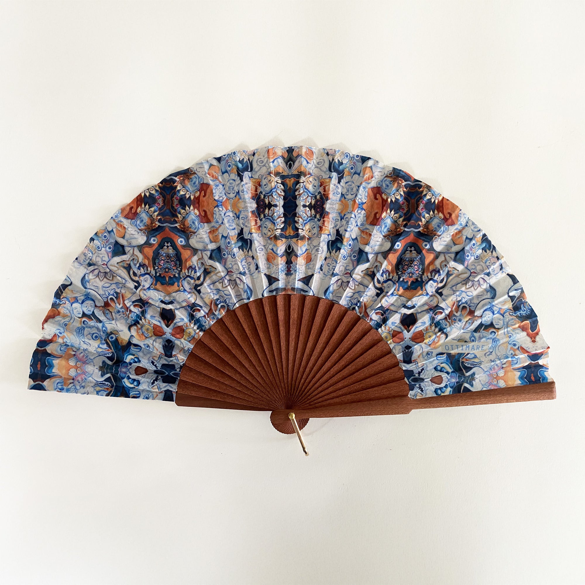 Elegante waaier van Ottimare met print op duurzame kunst zijde met Thangka dansers in donker blauw en sienna oranje. Het frame is van Kotibé hout en handgemaakt in Spanje. De waaier heeft een goudkleurige ring.